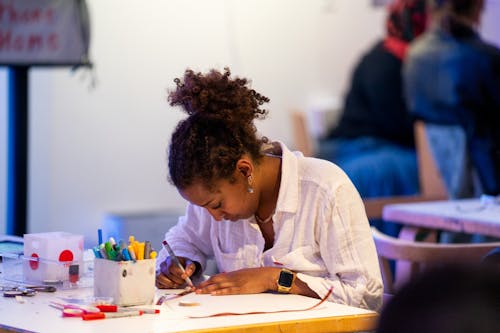 An adult with dark skin uses markers to make artwork while seated at a table.