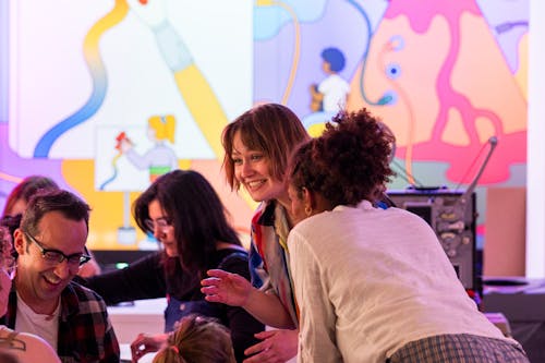 A group of adults laugh and talk to one another in a colorful artmaking room.