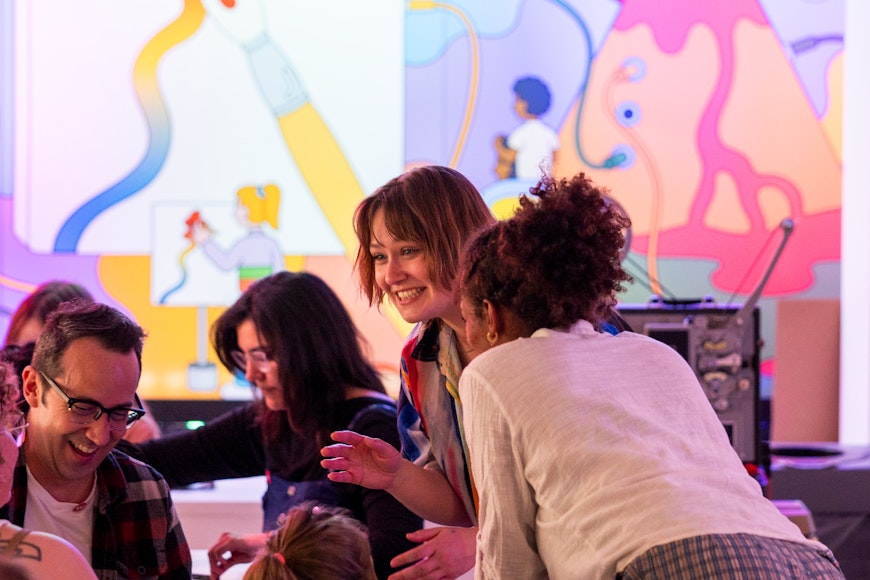 A group of adults laugh and talk to one another in a colorful artmaking room.