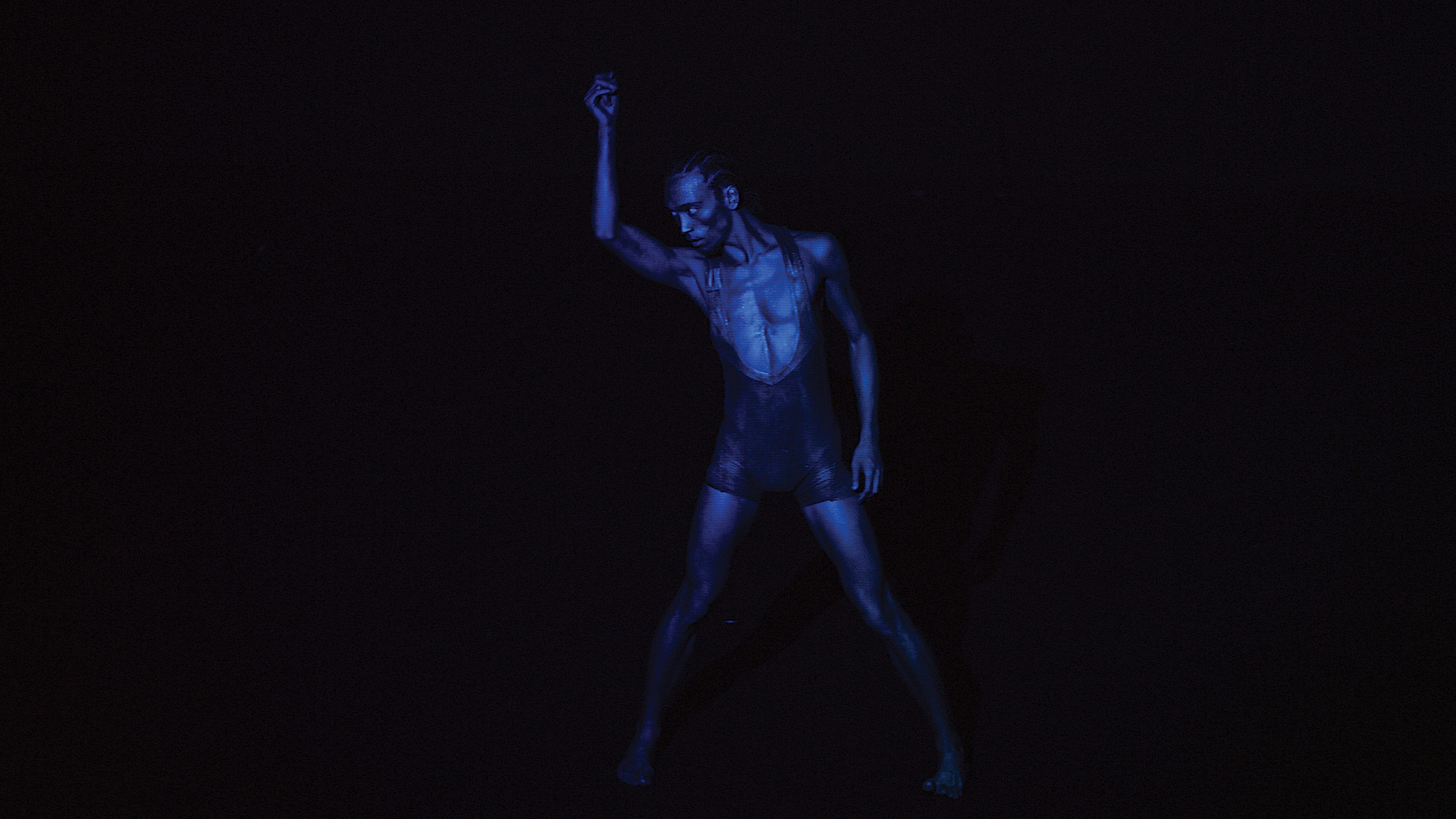 Against an all-black background, a blue monochromatic image of one dancers with dark skin holding their right hand above their head.