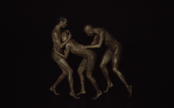 Against an all-black background, a monochromatic image of three dancers with dark skin who are entwined and holding each other.