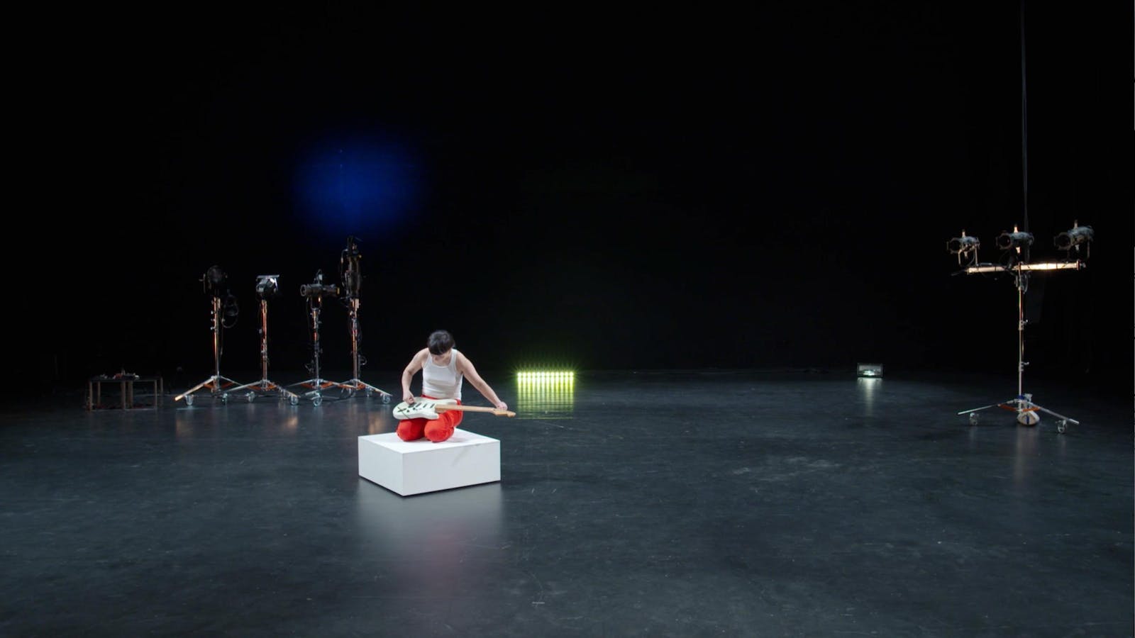 A dark theater stage with lighting tripods. A person sits on a small platform and plays a guitar on their lap.