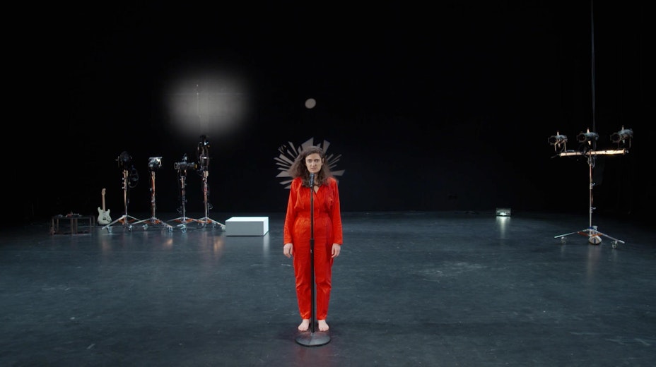 A dark theater stage with lighting tripods, a small platform and a guitar. A person in red stands front and center at a mic.