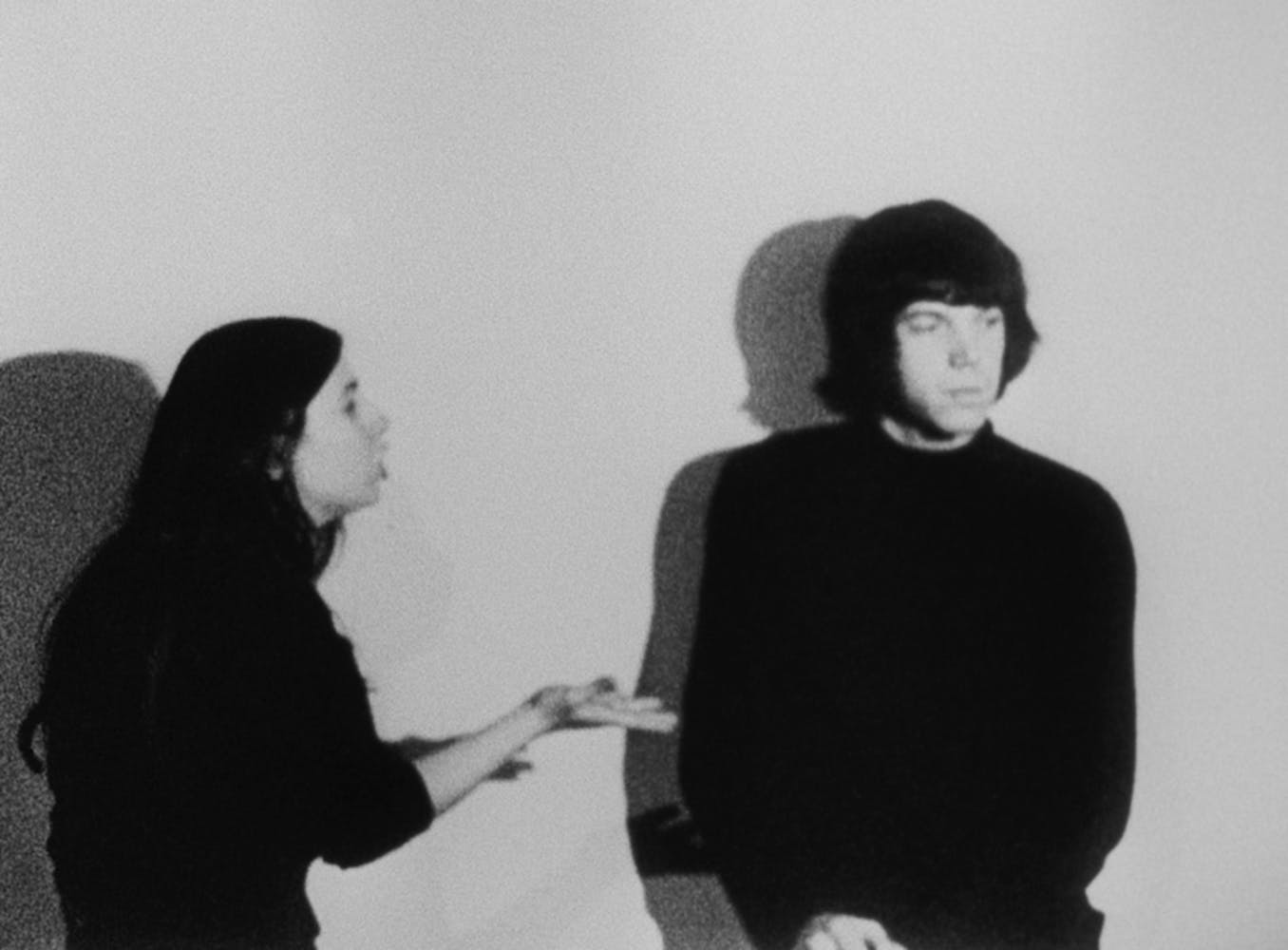 A black and white image of a couple standing against a white backdrop. The woman on the left addresses the man on the right, who is looking away from her.