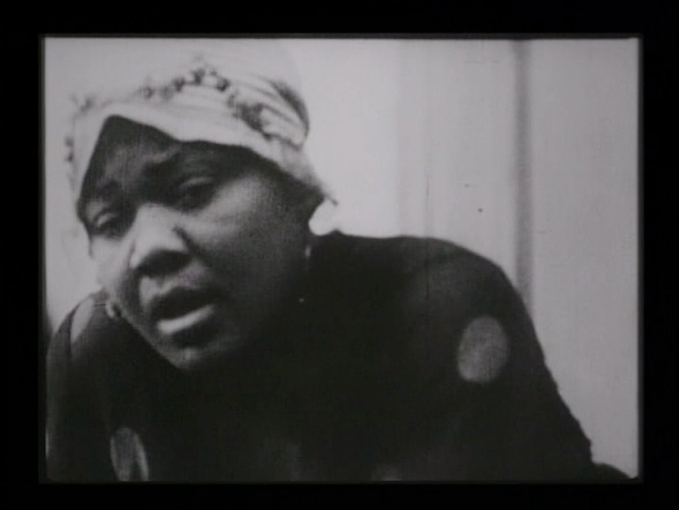 Black and white still of Bessie Smith singing. She leans to the left side of the image and wears a polka dot dress and a hat.