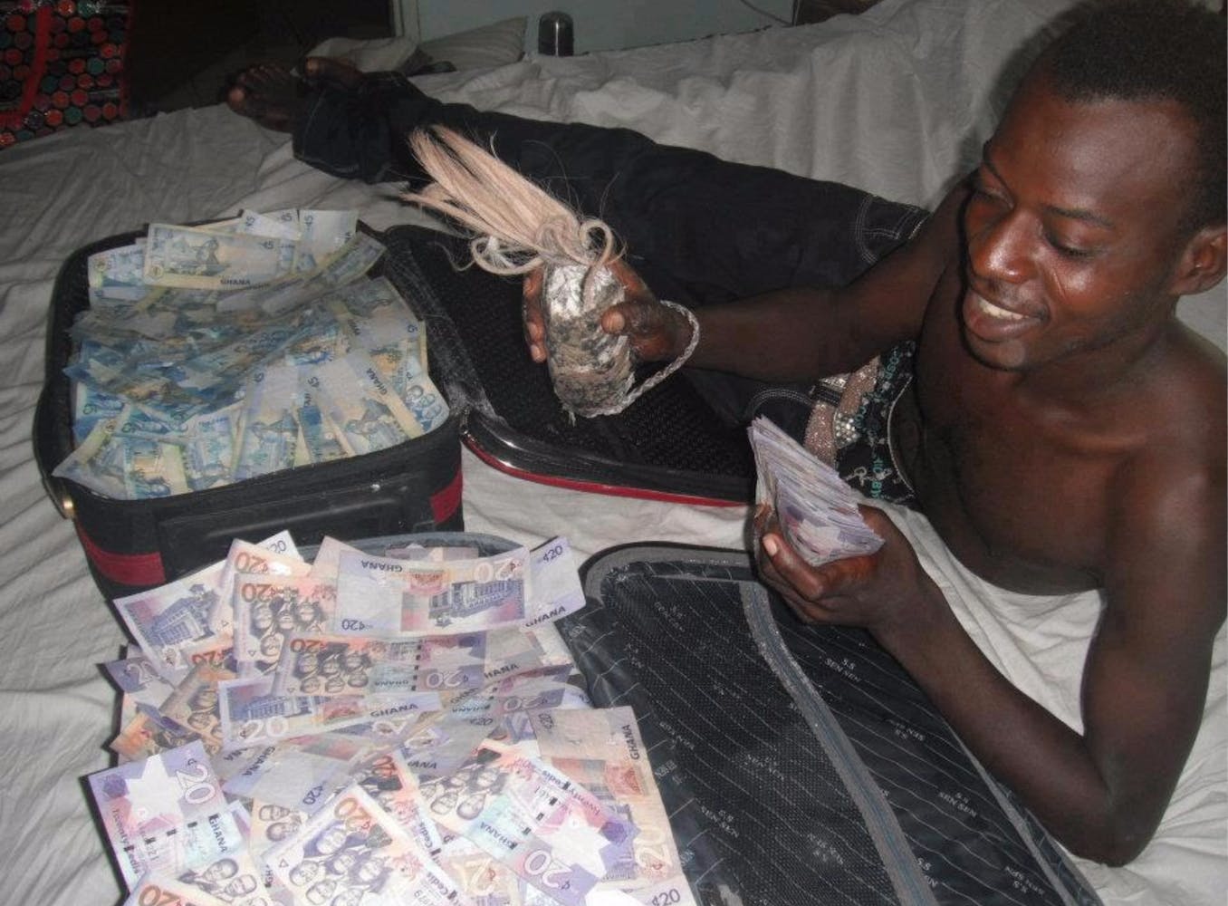 A shirtless young man lies on a bed in front of cases of cash with smiling and holding a talisman made from string, hair, and fabric.