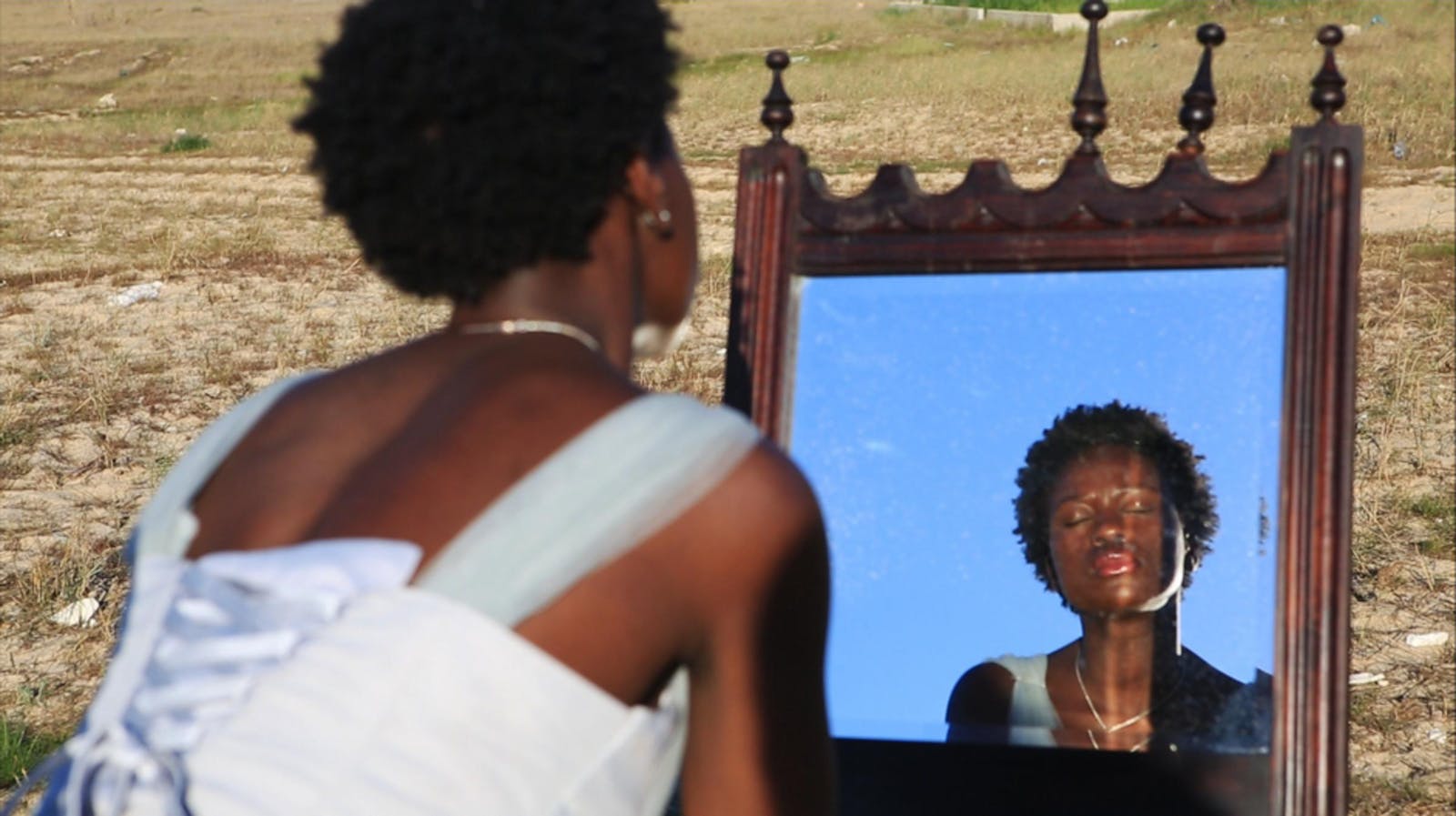 A woman with dark skin and short hair sits outdoors in front of a mirror with her eyes closed. Her back faces the camera.