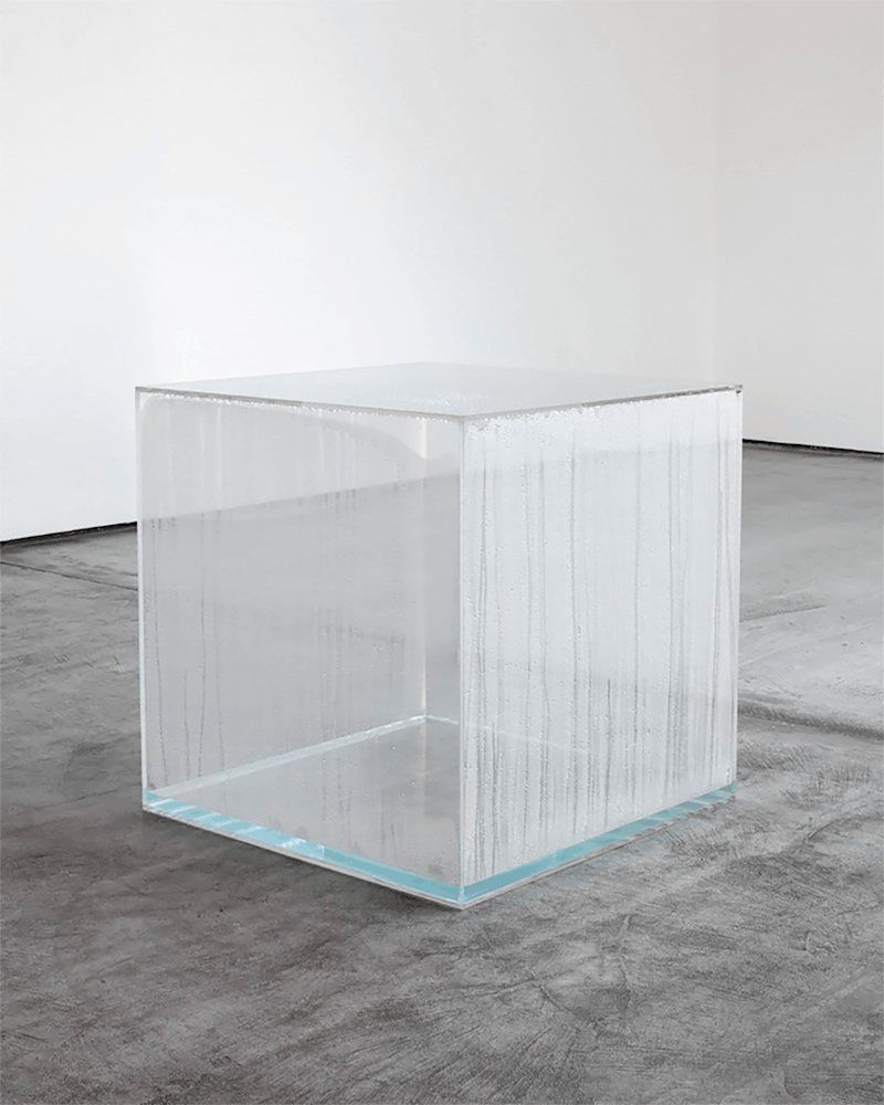 A large clear square with condensation inside sits on the floor of a white art gallery.