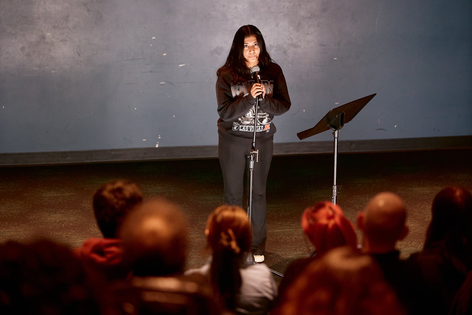 A teenager with dark long hair wearing a black sweatshirt speaks into a microphone on a stand in front of a crowd.