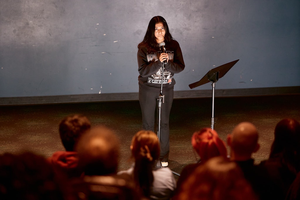 A teenager with dark long hair wearing a black sweatshirt speaks into a microphone on a stand in front of a crowd.