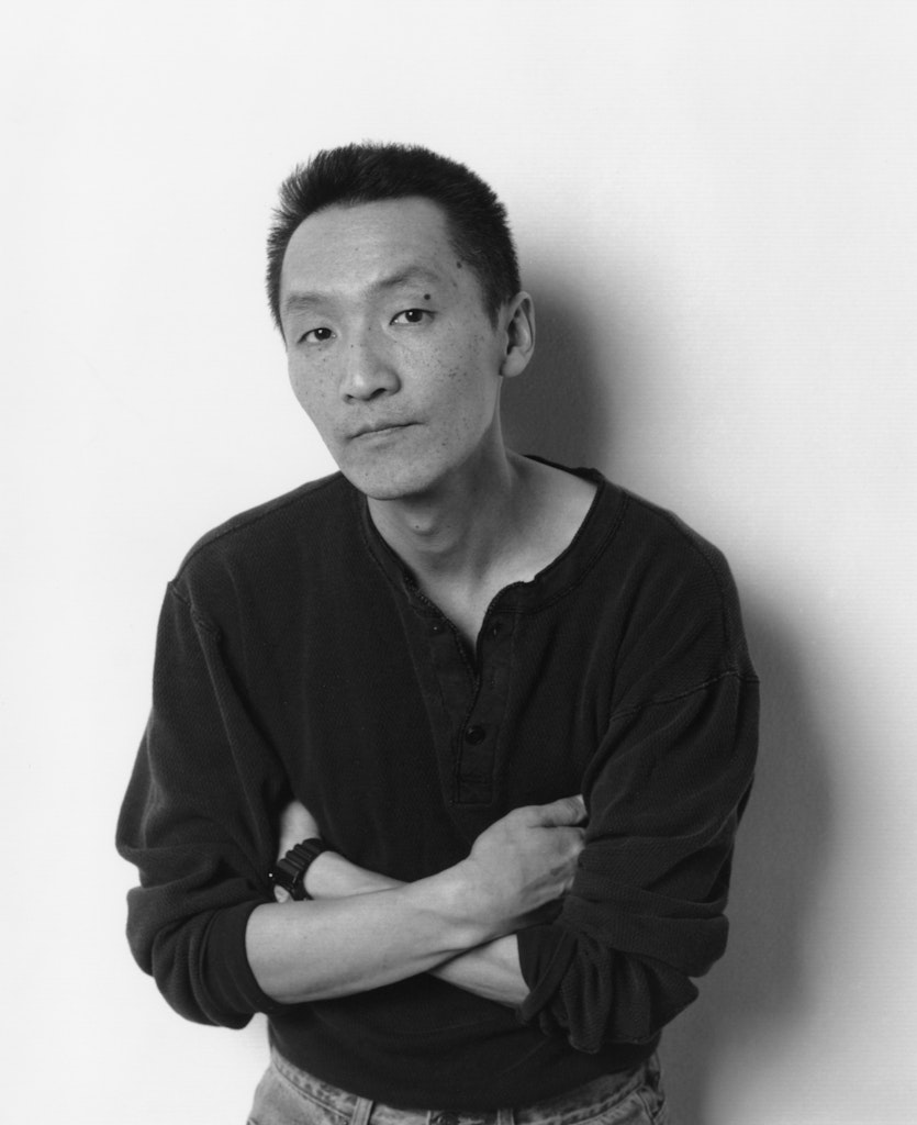 A black and white photo of an adult with meduim light skin wearing a black shirt and has short black hair looks at the viewer with arms crossed.