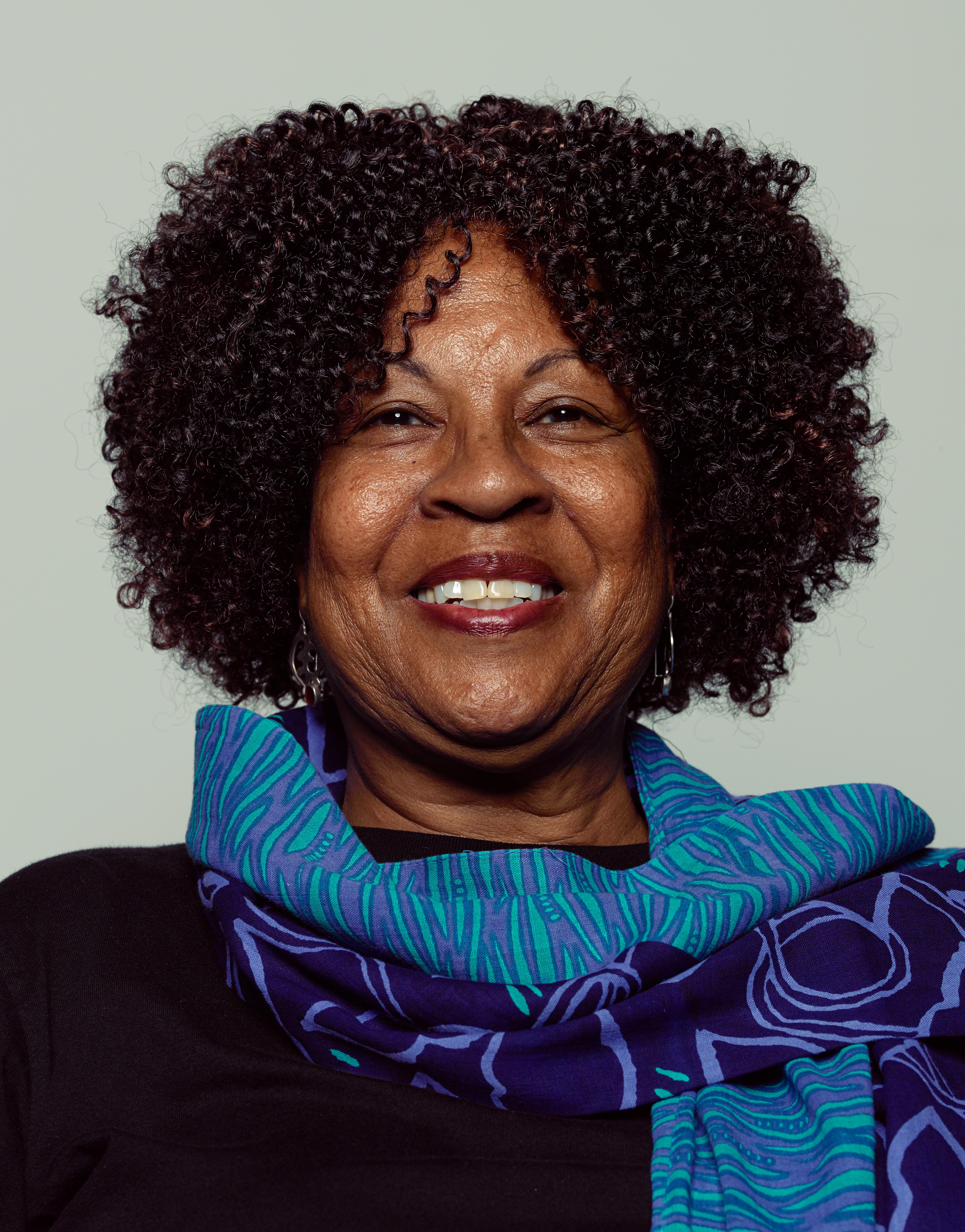 Portrait of an adult with dark skin and curly black hair wears a blue scarf and smiles at the viewer.