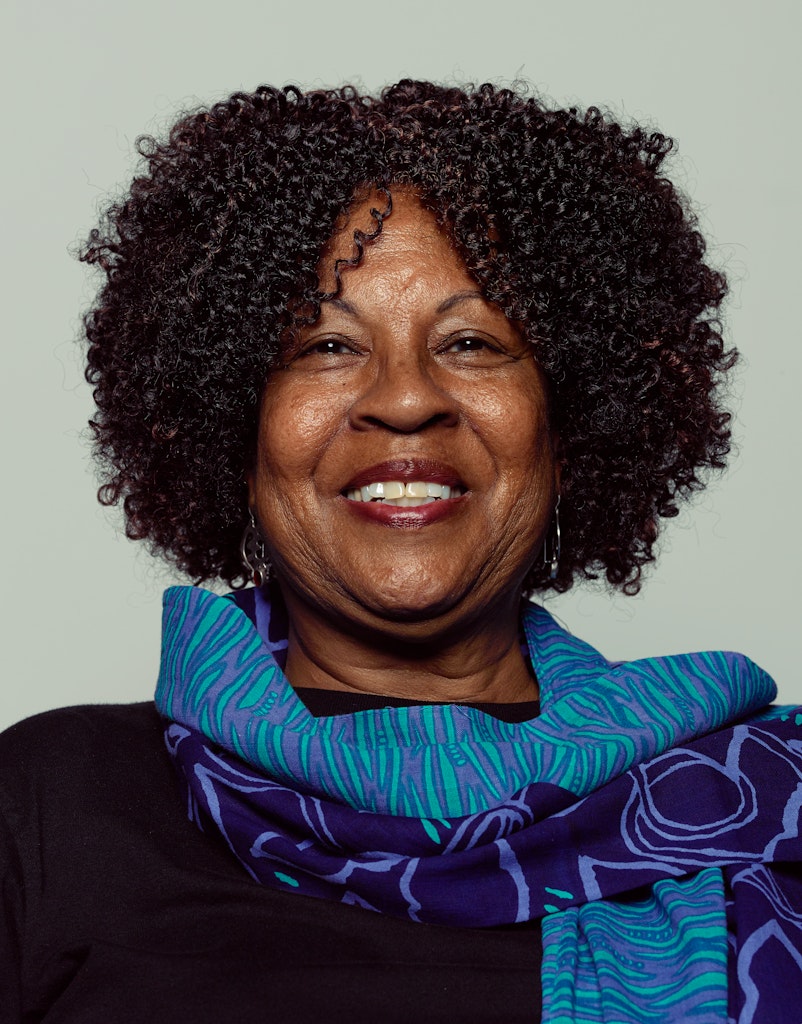 Portrait of an adult with dark skin and curly black hair wears a blue scarf and smiles at the viewer.