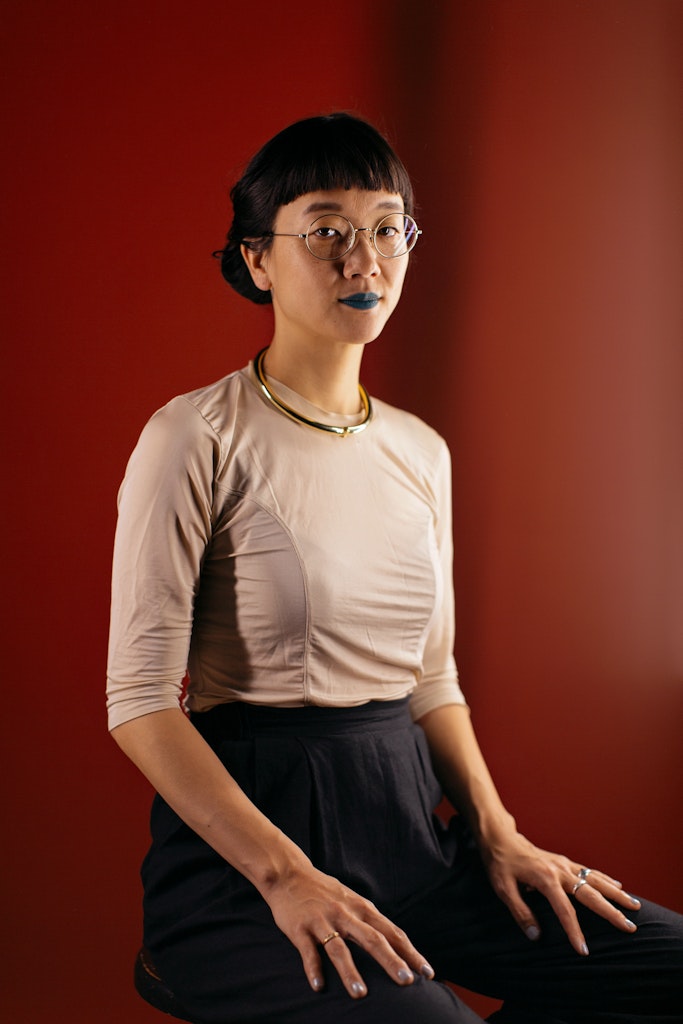 A woman with short dark hair and wearing glasses sits on a stool in front of a red background and looks at the viewer.