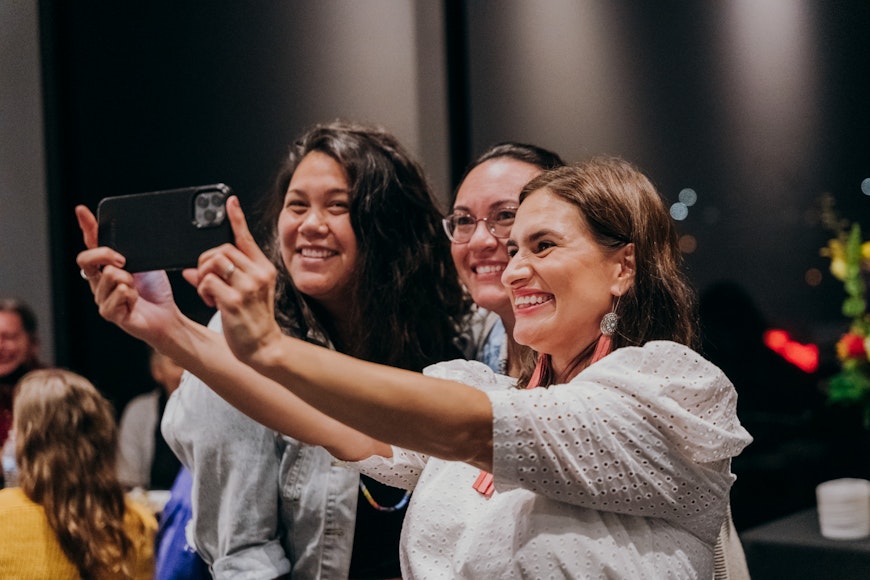 Three adults smile while one of them holds out a camera to photograpgh the group.