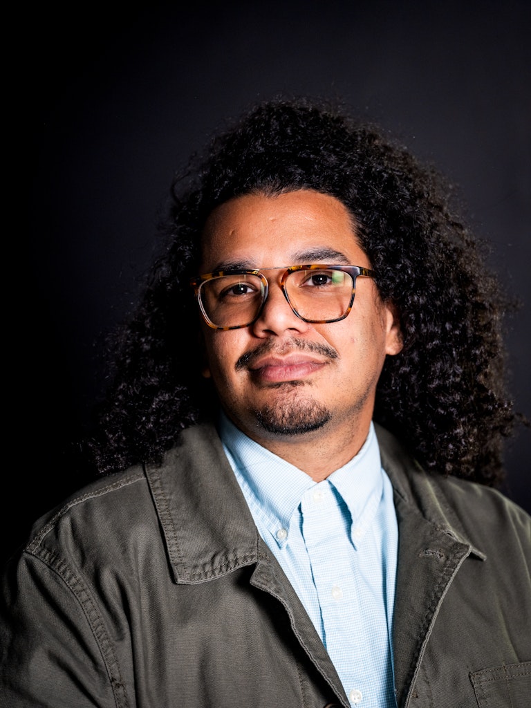 Portrait of an adult with medium brown skin, long curly black hair and tortious shell glasses, wearing a blue shirt and gray jacket looks at the viewer.