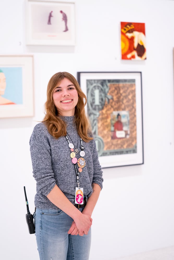 A woman folds her hands and smiles at the viewer while standing in front of artworks hanging on a wall in an art gallery.