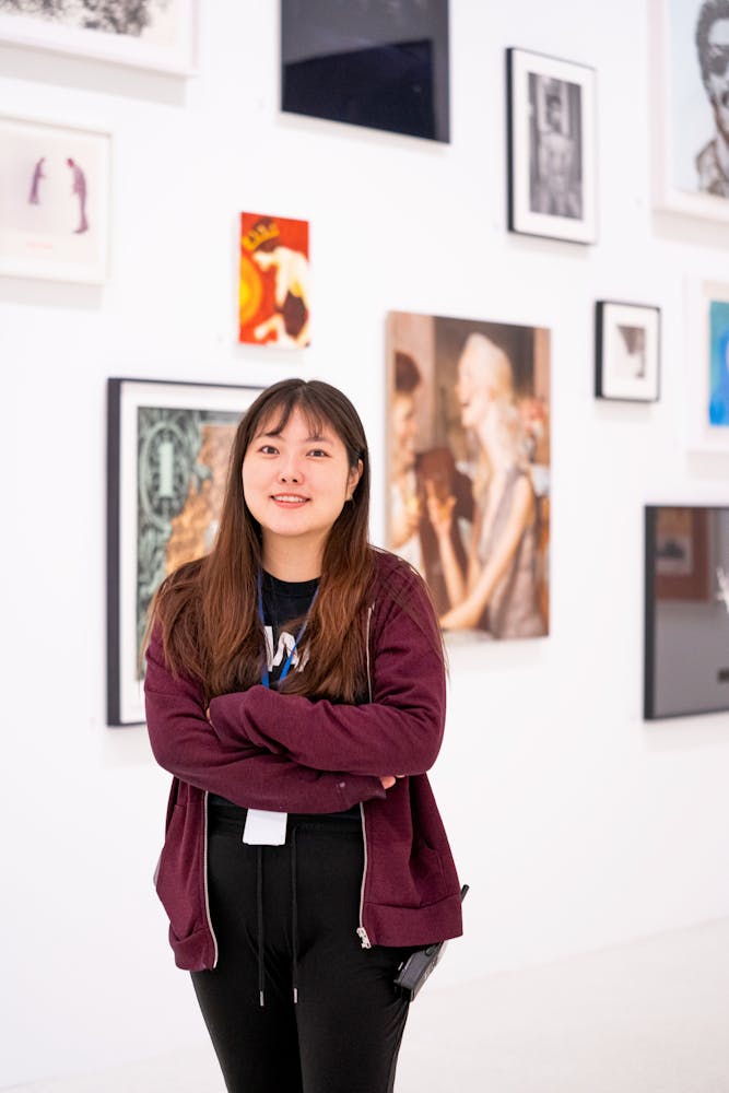 A woman folds her arms and smiles at the viewer while standing in front of artworks hanging on a wall in an art gallery.