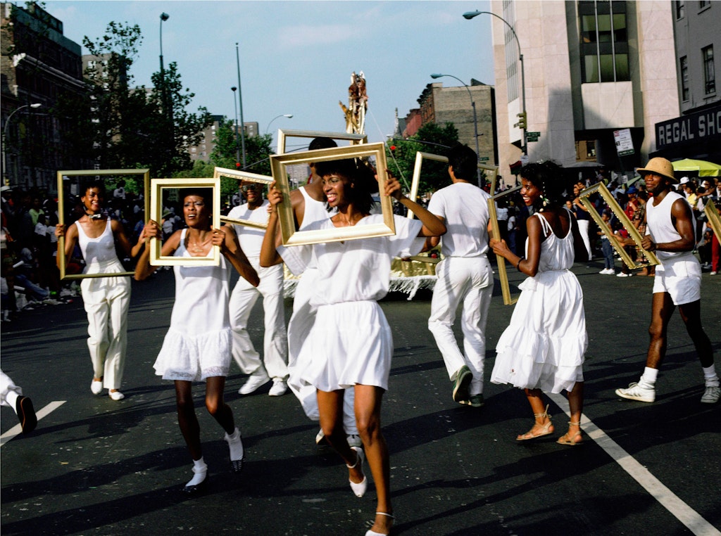 A group of people with dark skin and white clothing are walking in a street holding up gold frames over their faces.