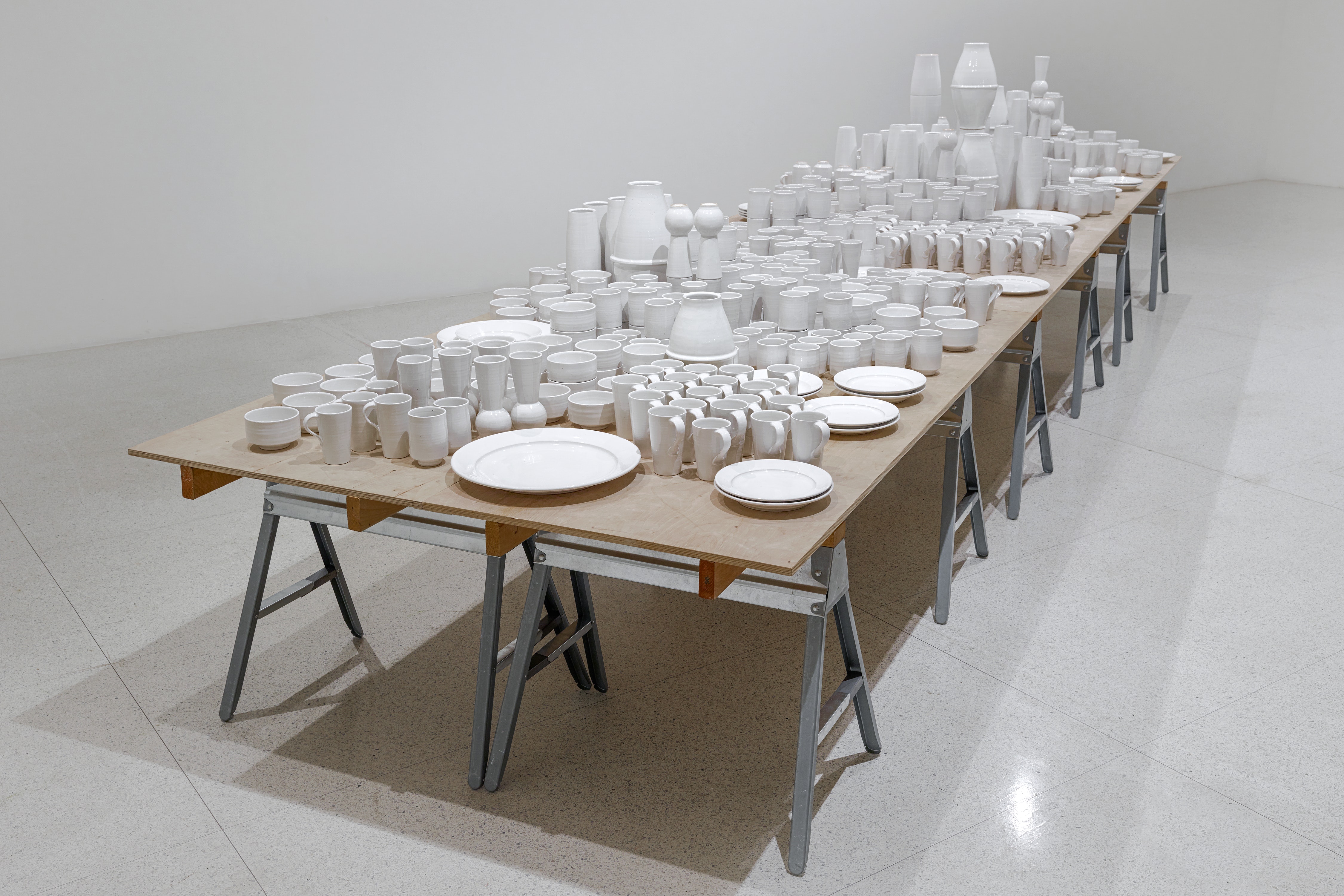 A variety of white cermaic cups, plates, bowls, and vases are collected on a wooden table with metal saw horse legs.