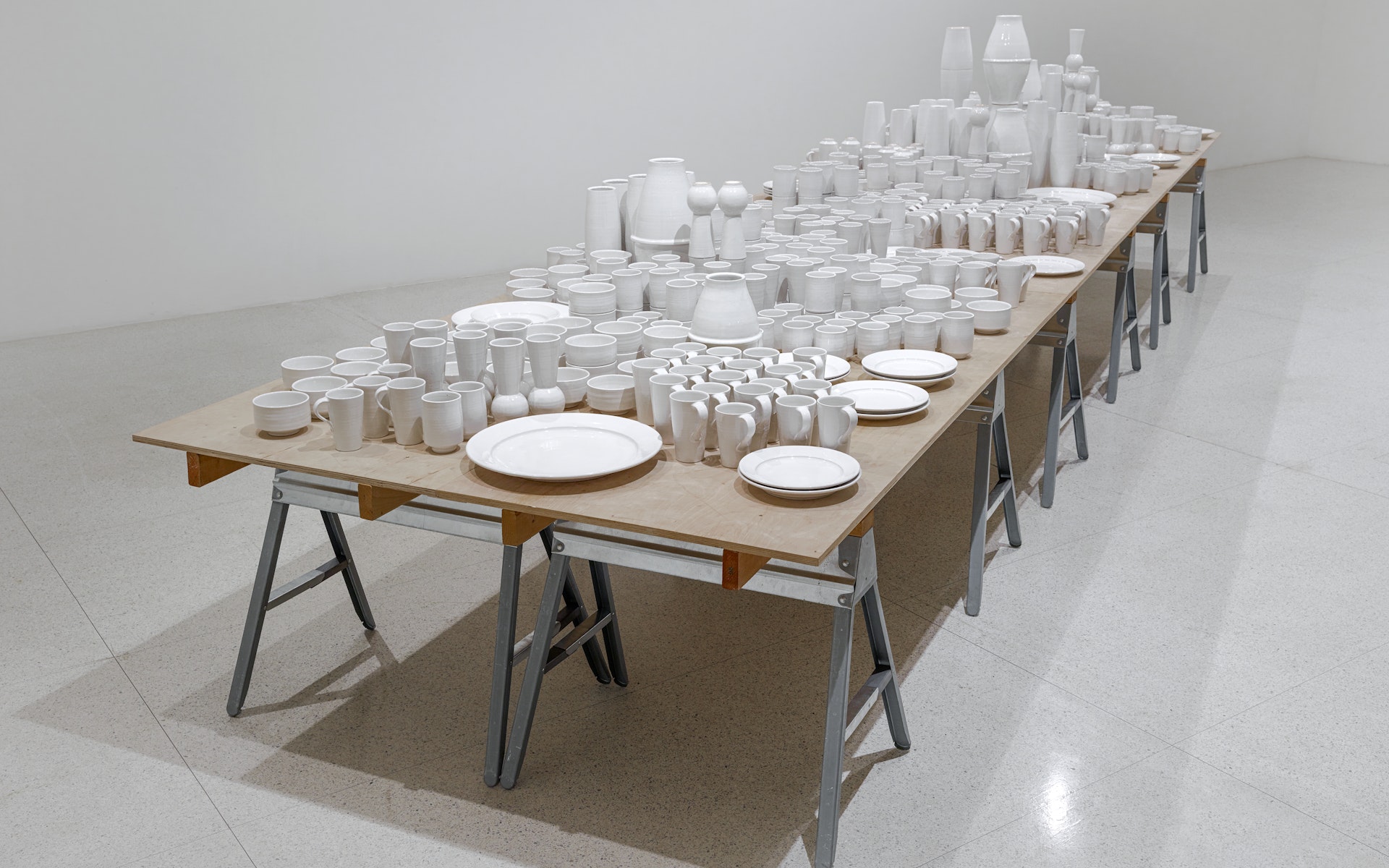 A variety of white cermaic cups, plates, bowls, and vases are collected on a wooden table with metal saw horse legs.