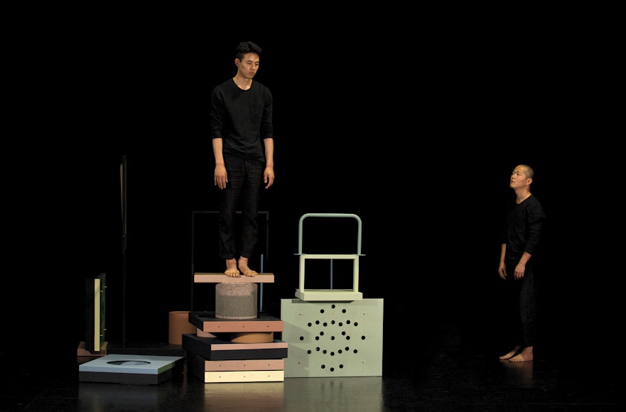 Black stage with two men in black clothes, one is standing on top of stacked objects