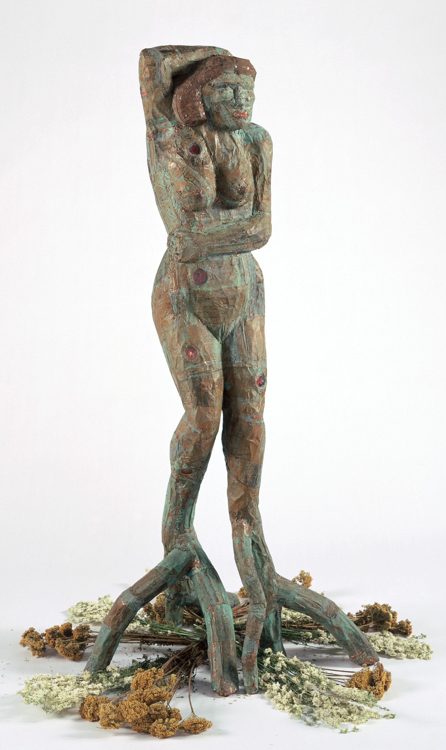A sculpture of a figure with tree legs and dried flowers beneath sits in a white space.
