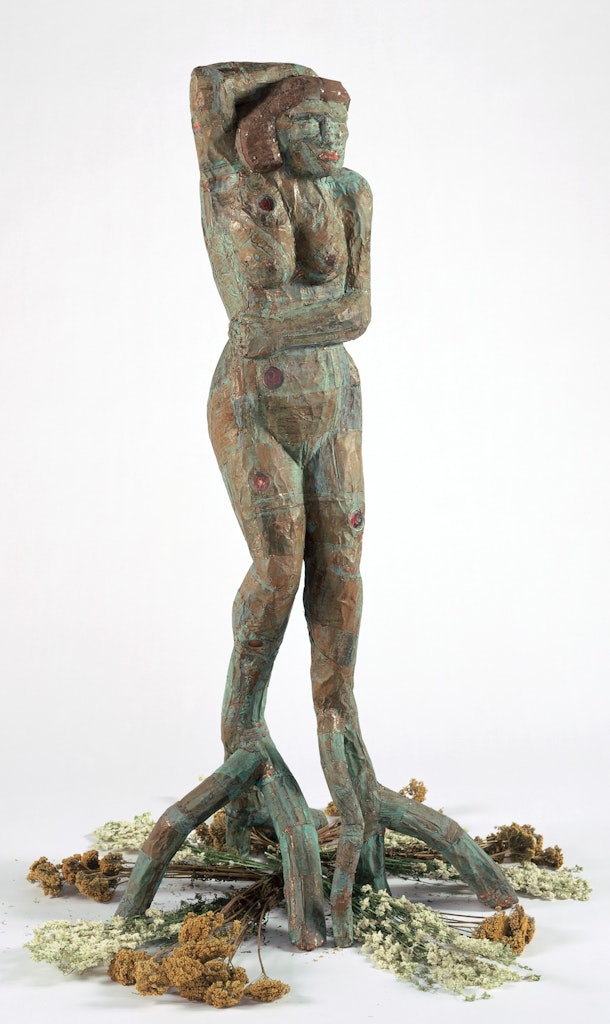 A sculpture of a figure with tree legs and dried flowers beneath sits in a white space.