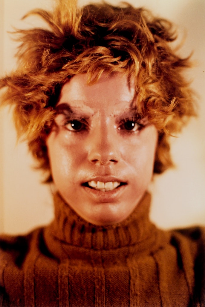 An adult with medium light skin faces the viewer with messy orange hair and a brown sweater.