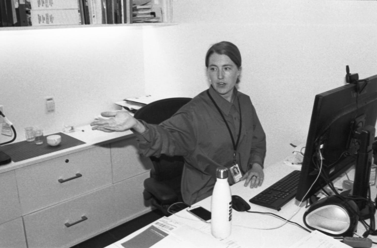 A woman sits in a desk in front of a computer while turning and gustering to someone off frame.