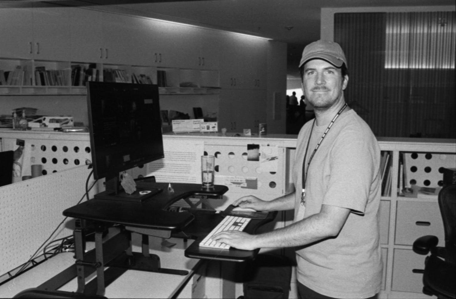 A man in a baseball hat stands and a stadnign office workspace and smiles at the viewer.
