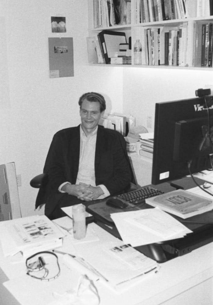 A man in a suite sits at a desk in an office and smiles at the viewer.