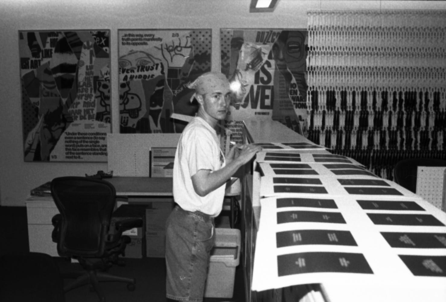 An adult with a bandana over their hair stands looks at the viewer while standing and examining printed documentsi n a museum office.