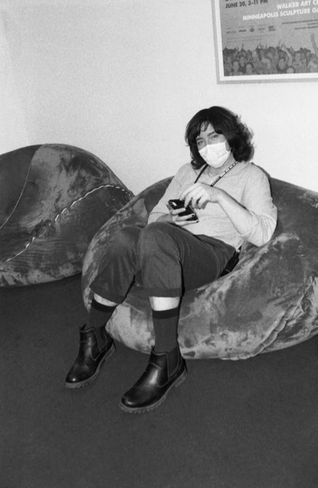 An adult wearing a face mask looks at the viewer while sitting on a beanbag and holding their cell phone.