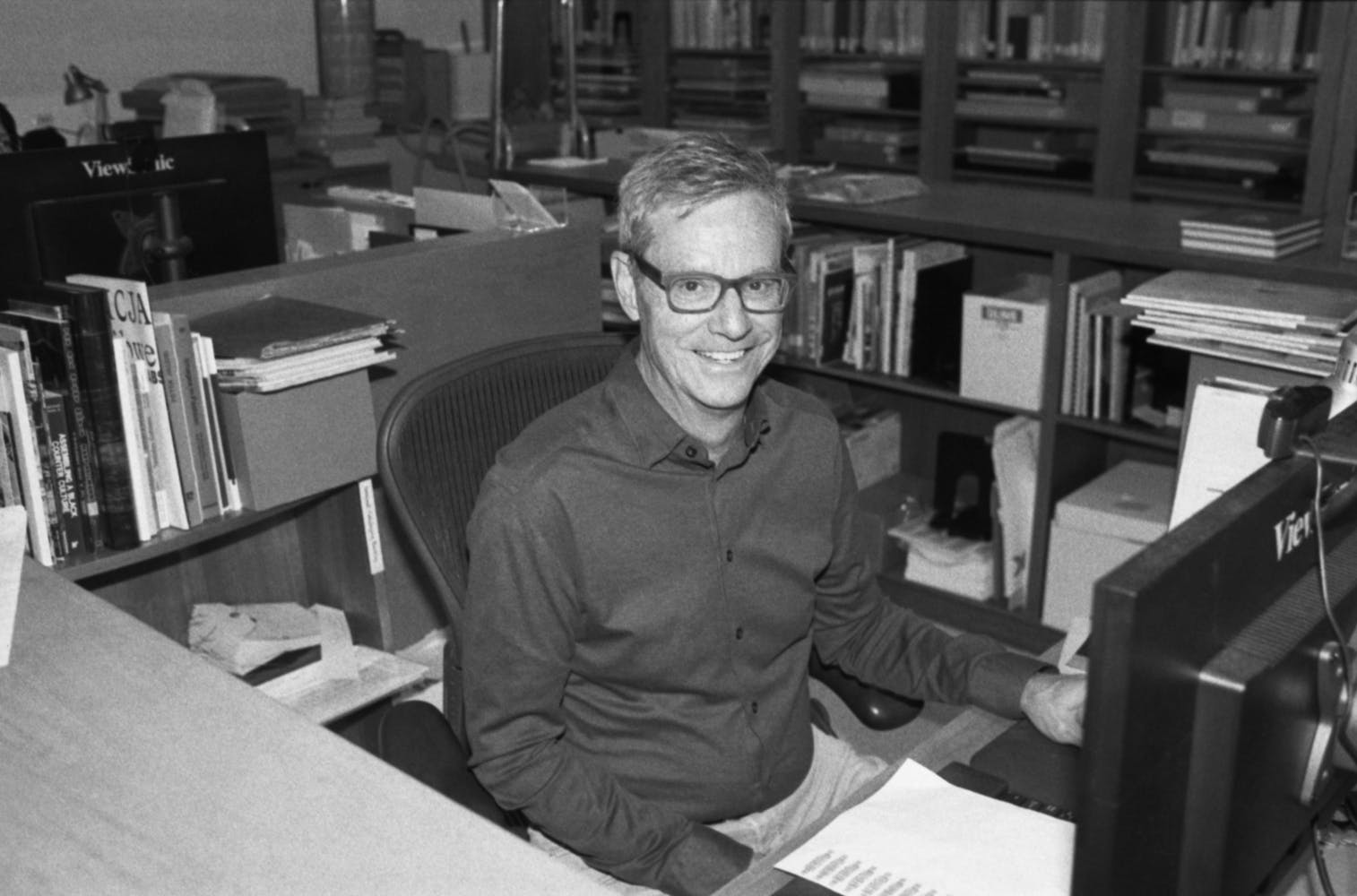 A man wearing glasses smiles while looking at the viewer as they sit at a desk among shelves of books in a library.