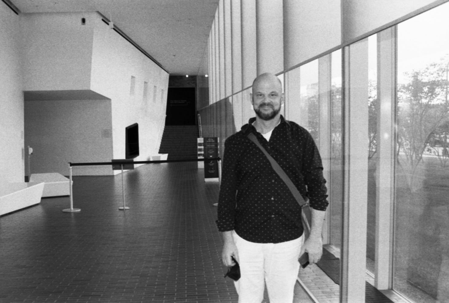 A man stands in a museum hallway smiling while lookinng at the viewer.