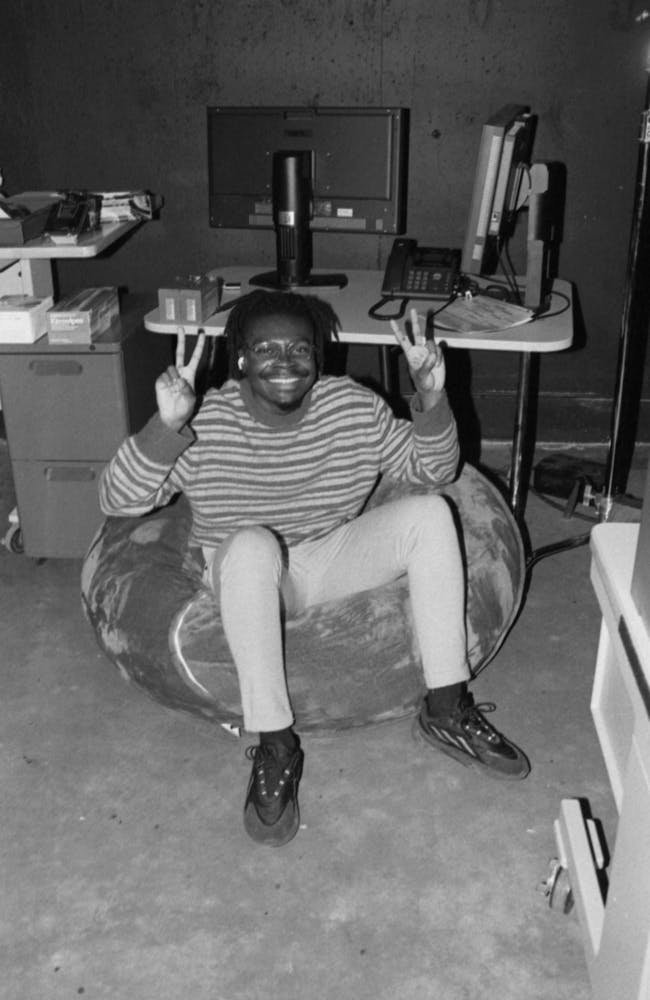 A man smiles at the viewer with holding up two peace signs with his hands as they sit on a beanbag in a photo studio.