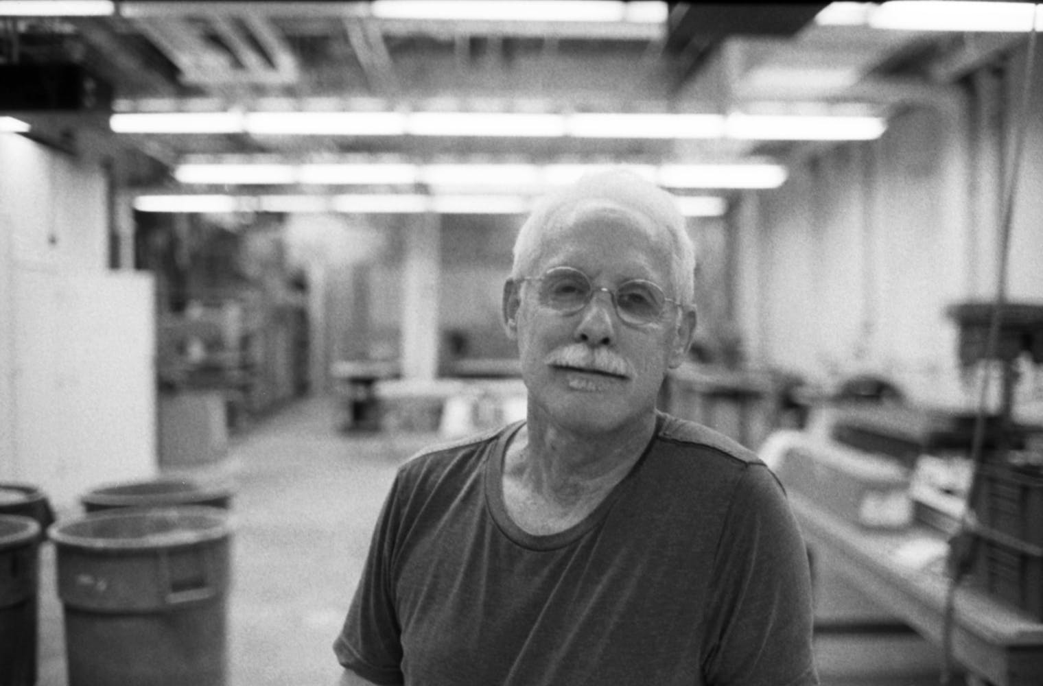 A man with glasses looks at the viewer while stanidng in a large woodworking shop.