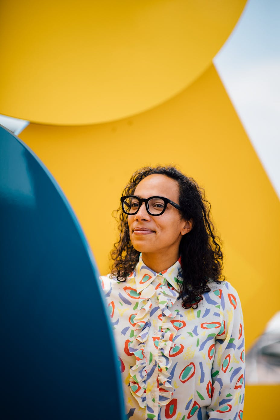 A woman wearing glasses stands smiling in a large metal yellow and blue sculpture.
