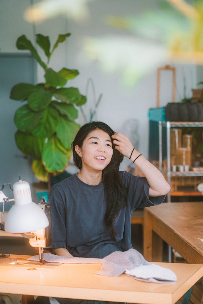 Through a window the back of a person with light skin and dark hair in a resperator and dark glasses welds together peices of metal in a studio is seen.