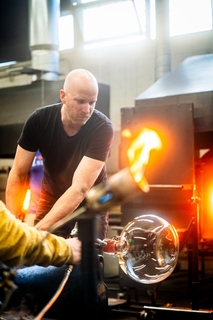 A light skinned adult in a black t-shirt is holding a glass blowing form mid action, a red hot flame is in foreground.