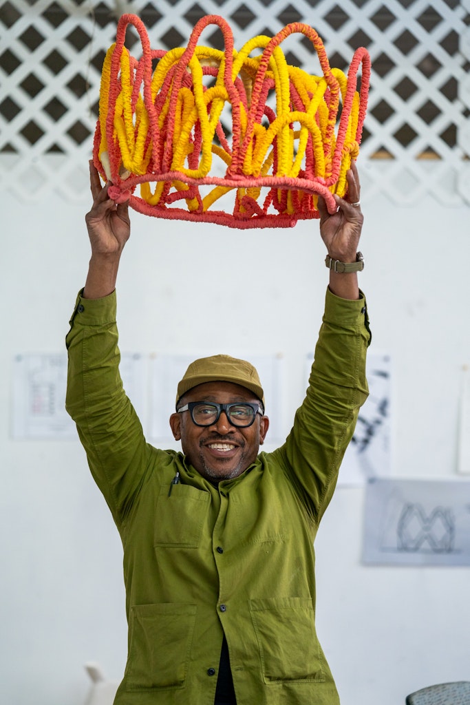 A dark skinned adult in a green shirt and hat stands behind colorful sculptures about to throw a sculpture up into the air.